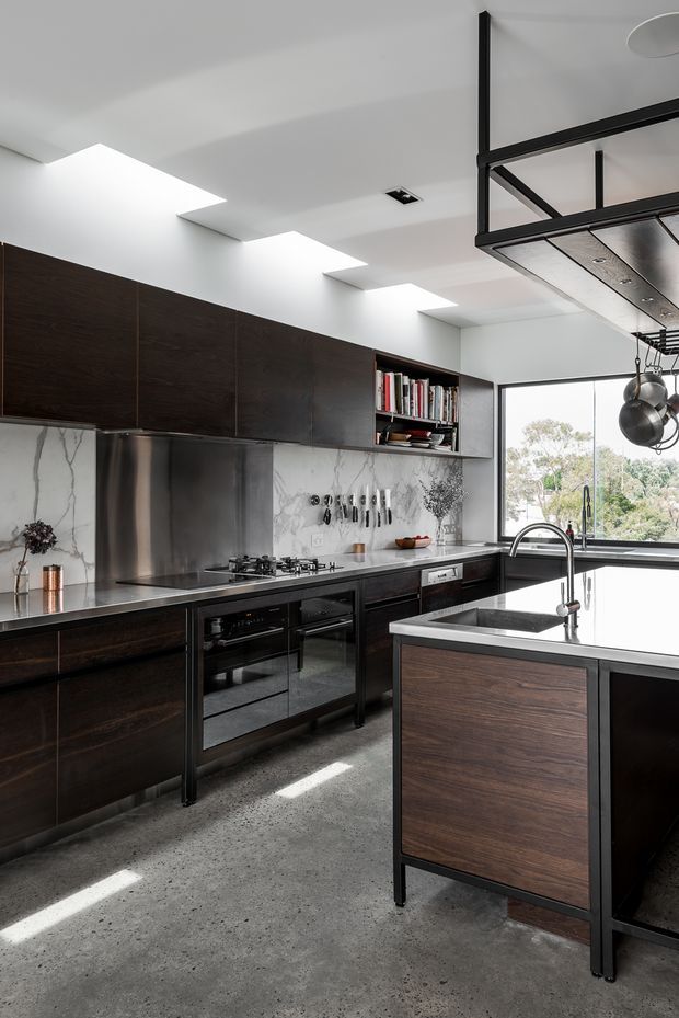 A sophisticated contemporary kitchen with dark timber cabinetry, stainless-steel work surfaces and appliances, a pale marble-look splashback, and a large central island. The kitchen features open shelving with books, hanging cookware, integrated lighting, expansive windows, and a polished industrial detailing palette. A concrete floor and black-framed overhead structure reinforce the modern industrial character.