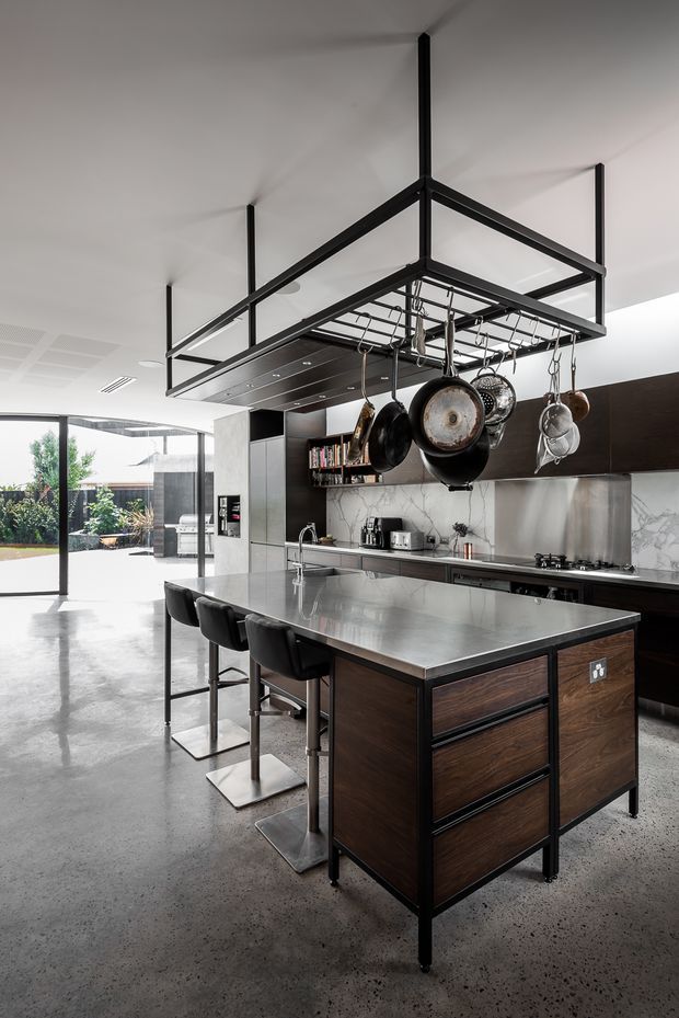 A spacious contemporary kitchen featuring a large stainless-steel island with dark timber cabinetry and seating for three. Black steel overhead shelving suspends cookware above the island, while the rear wall includes dark cabinetry, marble-look splashback, integrated appliances, and a long cooking bench. Polished concrete flooring and expansive glazing connect the kitchen to an outdoor garden area.