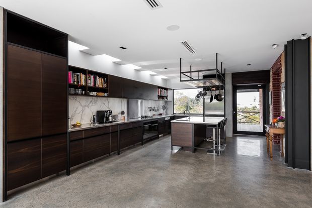 A spacious contemporary kitchen with dark timber cabinetry extending along the left wall, open shelving, a pale marble-look splashback, stainless-steel appliances, and a central island with bar stools. The room has a polished grey concrete floor, white ceiling with recessed linear lighting, exposed black metal hanging storage, and a brick feature wall beside full-height glazed doors. Natural light enters from the rear, creating a refined industrial atmosphere.