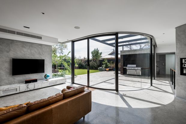A bright contemporary living room with polished concrete flooring, a large grey textured feature wall and built-in media console, and a brown leather sectional sofa. Full-height black-framed glazing with a curved corner opens the room visually to a landscaped courtyard and covered outdoor entertaining area with an outdoor kitchen. Strong sunlight casts geometric shadows across the interior.