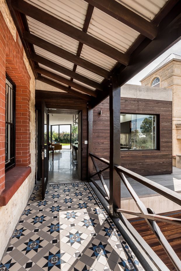 A covered outdoor circulation terrace or balcony links a historic brick-and-stone building with a contemporary timber-clad addition. The space has dark exposed timber beams supporting a translucent corrugated roof, dark timber and metal balustrades, and a patterned geometric encaustic-style tiled floor in grey, cream, black and blue. Glazed doors open into a bright interior, while a large window reveals a modern kitchen or living space. The composition contrasts heritage masonry with a contemporary extension.