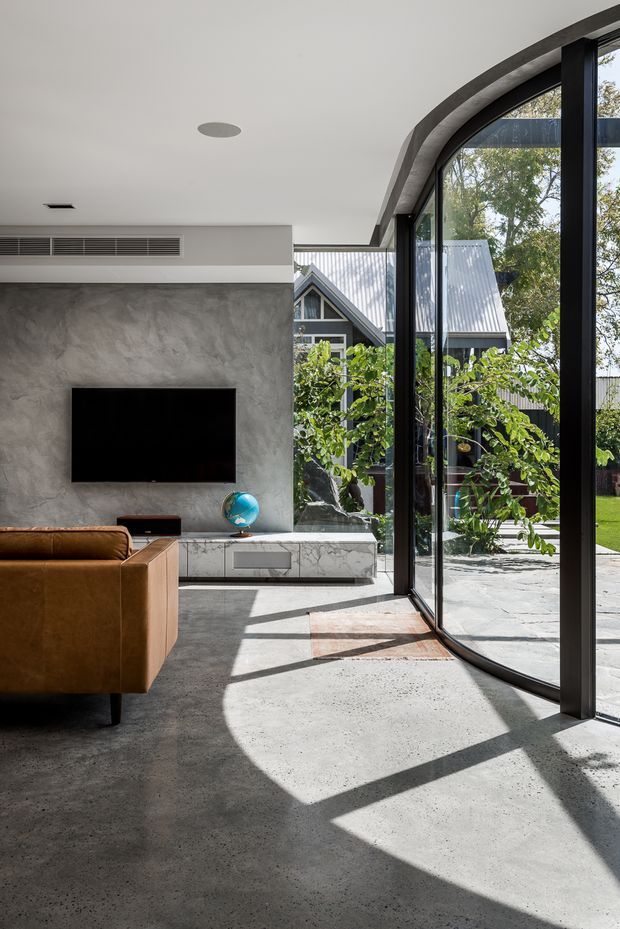 Bright contemporary living room with a polished concrete floor, textured grey feature wall, wall-mounted television, low marble-look media console, and tan leather sofa. A large curved floor-to-ceiling glazed corner opens toward a landscaped courtyard and reveals a pitched-roof building outside. Strong sunlight casts geometric shadows across the floor.