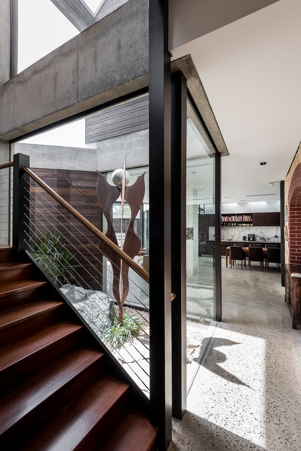 Interior view of a contemporary multi-level home featuring a dark timber staircase with a slim black metal and cable balustrade, exposed concrete walls and beams, extensive black-framed glazing, and a polished terrazzo floor. A bright kitchen and dining area with dark cabinetry and open shelving is visible beyond the glazed partition, while an internal courtyard or lightwell brings daylight into the stair hall.