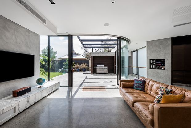 A bright contemporary living room opening onto an outdoor entertaining area. The room features a large television mounted on a grey concrete-effect feature wall, white marble-look built-in media cabinetry, polished concrete flooring, a large cognac leather sofa, and extensive floor-to-ceiling glazing with a curved glass corner. Outside, a covered patio with an outdoor kitchen or barbecue is visible.