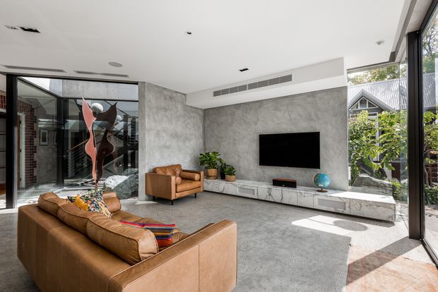 Bright contemporary living room with a large tan leather sectional sofa and matching armchair, polished grey concrete-style walls and floor, a floating stone-look media console, wall-mounted television, indoor plants, and expansive black-framed glazing. A sculptural artwork and courtyard greenery are visible through the glazing, with strong natural light creating an airy, refined atmosphere.