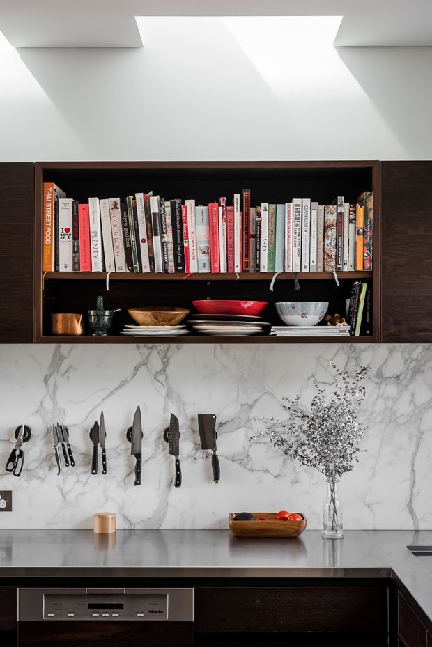 A refined contemporary kitchen featuring dark timber cabinetry, an open display shelf filled with cookbooks and ceramics, a dramatic white marble backsplash with grey veining, and a sleek grey stone benchtop. Knives and utensils are mounted beneath the shelf, while a built-in stainless-steel dishwasher sits below the counter. A large recessed ceiling opening provides bright natural light.