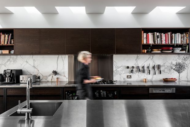 Contemporary kitchen with a large dark island, dark timber cabinetry, white marble-look splashback, integrated appliances, open shelving, and a dramatic skylit ceiling. A blurred person is moving through the space.