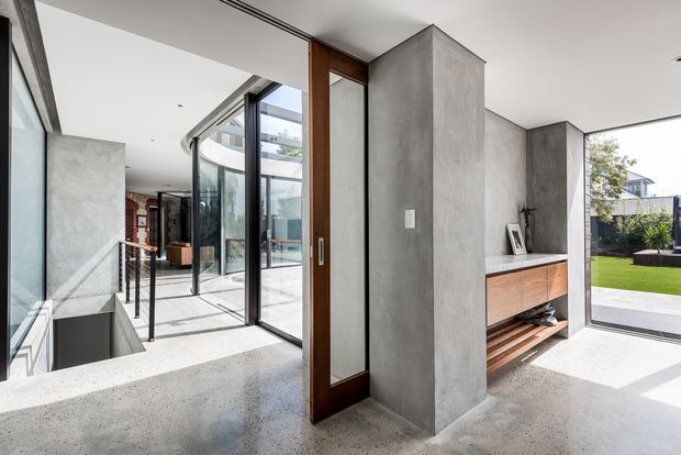 Bright contemporary open-plan interior with polished light terrazzo or concrete-style flooring, raw grey concrete-look walls and a central built-in media or storage joinery unit in warm timber. Floor-to-ceiling black-framed glazing and timber-framed doors open toward a landscaped courtyard and lawn, while a glazed internal walkway connects to an adjoining living area. The space has a clean, minimalist architectural character with strong natural light and indoor-outdoor flow.