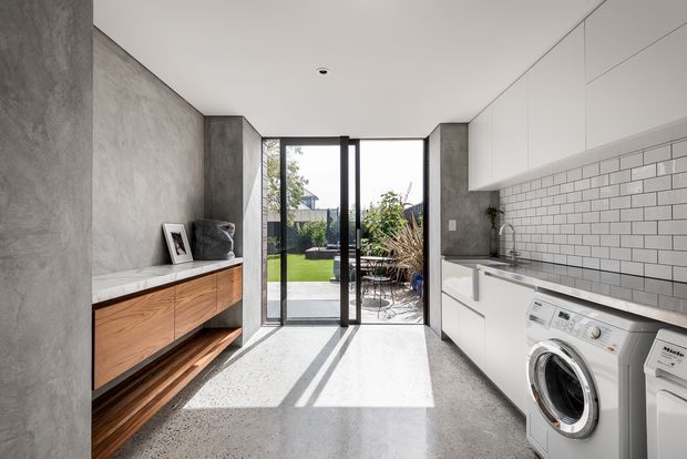Bright contemporary laundry with a long wall of white built-in cabinetry, integrated washing appliances, a stainless sink, and a white subway-tile splashback. The room has polished concrete flooring, a textured grey concrete-look wall, warm timber drawers and open shelving, and full-height glazed doors opening to a landscaped outdoor area.