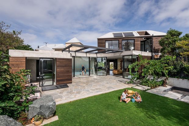 A contemporary multi-level residential home viewed from its landscaped rear courtyard. The house combines warm horizontal timber cladding, concrete walls, extensive floor-to-ceiling glazing, and a broad glazed connector beneath a dark metal-framed roof canopy. Solar panels are visible on the upper roof. A large paved stone terrace leads to a manicured lawn bordered by dense planting, raised concrete planters, and feature boulders.