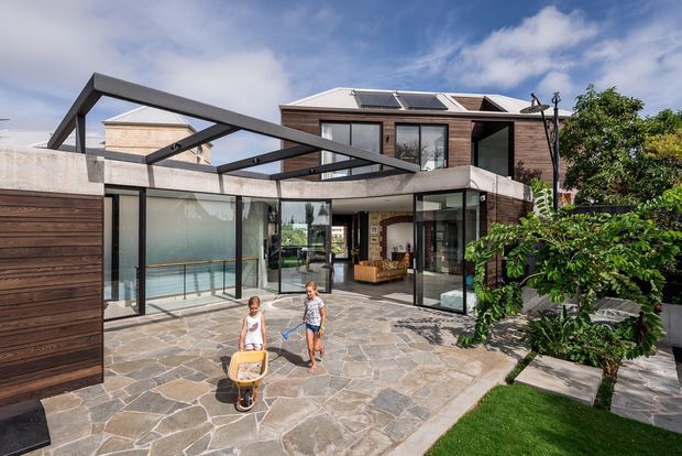 Contemporary multi-level residence with a sculptural steel pergola and exposed roof structure over a large paved courtyard. The home combines dark timber cladding, concrete forms, extensive black-framed glazing, and open-plan indoor-outdoor connections. A glazed living area and entry are visible, with landscaped planting and lawn around the stone-paved outdoor space.