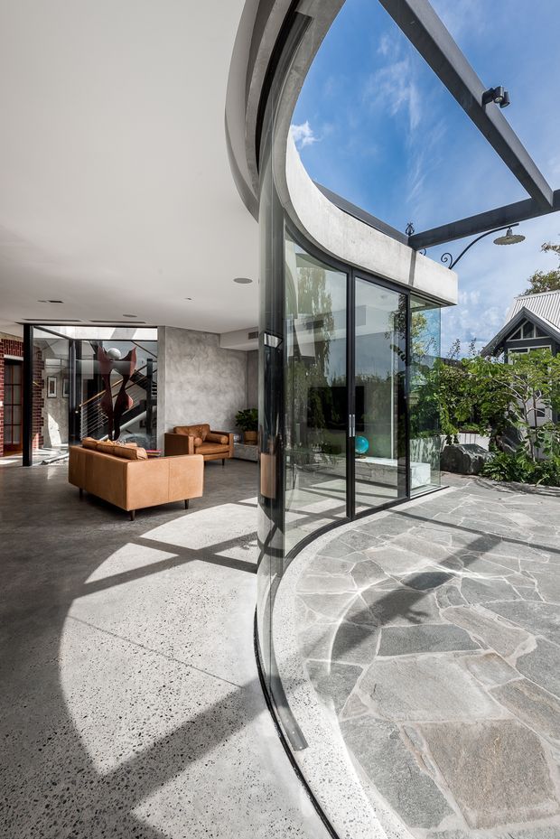 A contemporary indoor-outdoor living space with a dramatic curved glass wall and black-framed glazing opening onto a paved courtyard. The interior features polished concrete flooring, a white ceiling, exposed brick, textured grey wall finishes, and tan leather lounge furniture. A large overhead pergola structure and outdoor shower are visible above the irregular stone paving, with neighbouring architecture and greenery beyond.