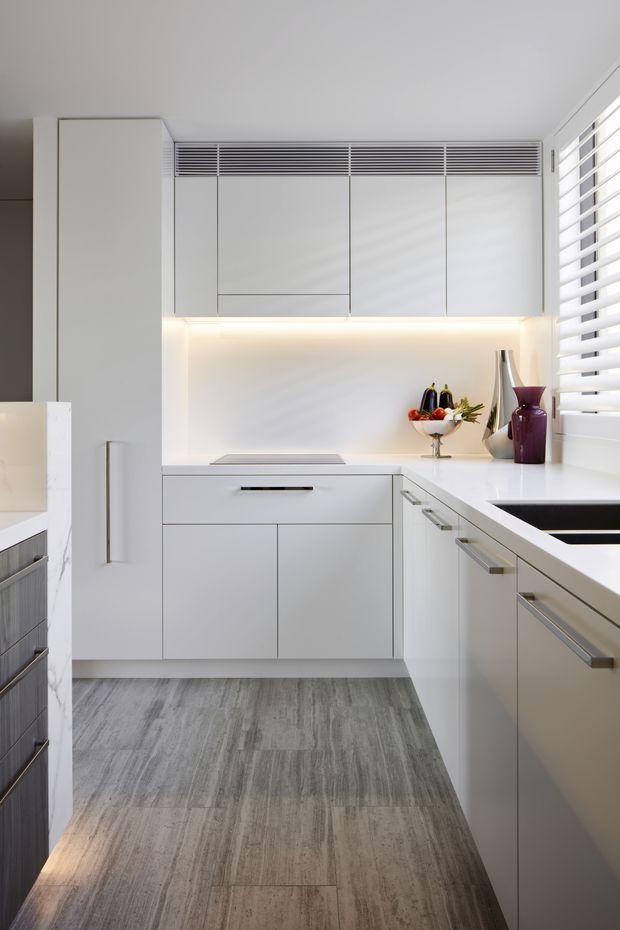 Bright, compact contemporary kitchen with extensive white handleless cabinetry, integrated upper ventilation grille, pale stone benchtops, under-cabinet lighting, dark inset cooktop, and grey stone-look flooring. White plantation shutters provide filtered natural light, while a small arrangement of decorative vessels and fruit adds colour.