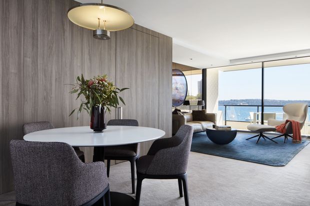 A refined open-plan dining and living area with a round white dining table and upholstered charcoal dining chairs in the foreground. A full-height timber-look feature wall divides the dining zone from the lounge, which contains a sculptural coffee table, lounge seating, a blue area rug, and a mid-century-style lounge chair. Floor-to-ceiling glazing opens to a bright coastal view, while a large circular pendant light anchors the dining area.