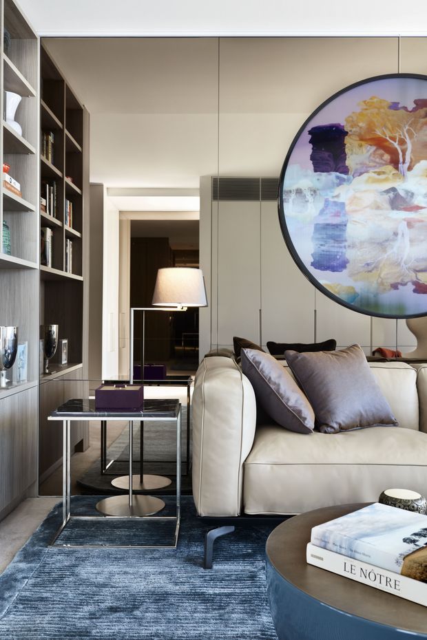 A refined living room with a cream upholstered sofa, grey-lilac cushions, dark timber built-in shelving, pale panelled cabinetry, a glass-and-metal side table, and a large round abstract artwork. A floor lamp and dark blue textured rug add layered, sophisticated detailing.