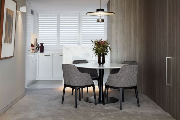Contemporary dining area with a round marble-look table, four upholstered dining chairs, a sculptural pendant light, and a large floral arrangement. White plantation shutters and white built-in cabinetry sit at the rear, while a dark wood-toned full-height wall or concealed joinery panel adds contrast. The room has a polished, refined, neutral palette with concrete-look flooring and soft grey furnishings.