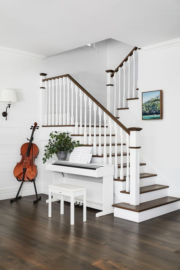 Bright, refined interior view of a traditional-style staircase and compact piano area. White painted walls, decorative cornices, turned white balusters, and dark timber stair treads and handrail create a crisp contrast. A white upright digital piano with sheet music and bench sits beneath the stairs, accompanied by a cello, plant, and wall-mounted lamp. Dark timber flooring runs throughout the space.
