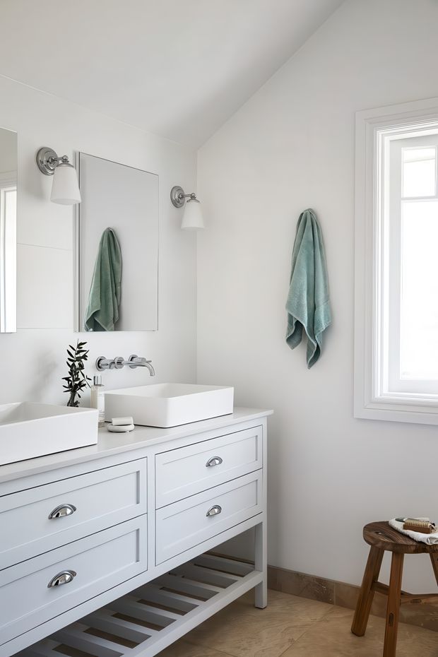 Bright, airy bathroom featuring a white double vanity with shaker-style drawers and chrome cup pulls, two white vessel basins, wall-mounted chrome faucets, rectangular mirrors, and chrome wall sconces. A pale aqua towel hangs beside a white-trimmed window, while the floor has a warm light wood-look finish and a small wooden stool sits near the window.