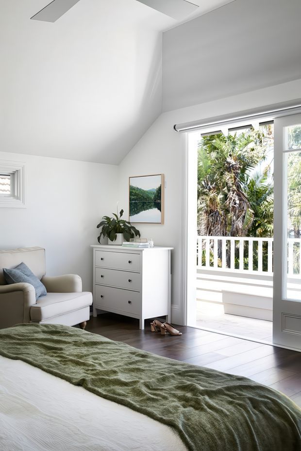 Bright, serene bedroom with white walls and a steeply angled ceiling, dark timber flooring, and a large bed dressed in white linens with a textured sage-green throw. A pale upholstered armchair, white chest of drawers, indoor plant, framed landscape artwork, and books furnish the room. Open glazed doors lead to a covered balcony or terrace with white balustrades and lush tropical planting outside.