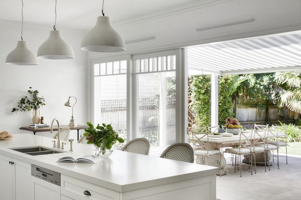 Bright white kitchen with a large central island, pendant lights, and glass doors opening to a covered outdoor dining terrace. The space features white shaker-style cabinetry, a pale benchtop, traditional trim, abundant natural light, and greenery visible outside.