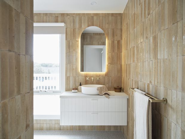 A compact, bright bathroom featuring full-height warm beige textured wall tiles, a floating white fluted vanity with a round vessel basin, and an arched backlit mirror. A window provides natural light, while brushed brass tapware and towel hardware add a warm accent. The floor is finished in a small-speckled grey material.