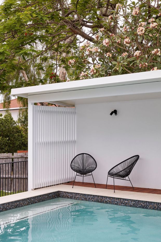 A compact modern outdoor pool courtyard with a pale blue tiled swimming pool in the foreground, bordered by a dark mosaic-tile edge and timber decking. A white flat-roofed pavilion or pool cabana contains two black woven lounge chairs, a black wall light, and a vertical white slatted privacy screen. Mature leafy and flowering vegetation surrounds the space, with glimpses of neighbouring residential structures beyond.