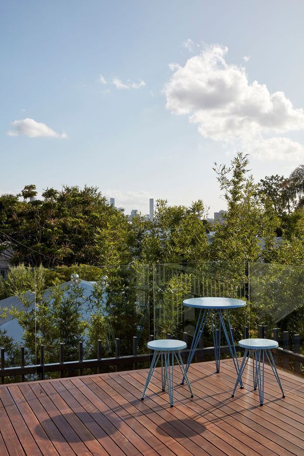 Sunny elevated timber deck with a small blue outdoor table and stools, enclosed by clear glass balustrading and surrounded by dense greenery. A distant urban skyline is visible through the trees beneath a bright blue sky.
