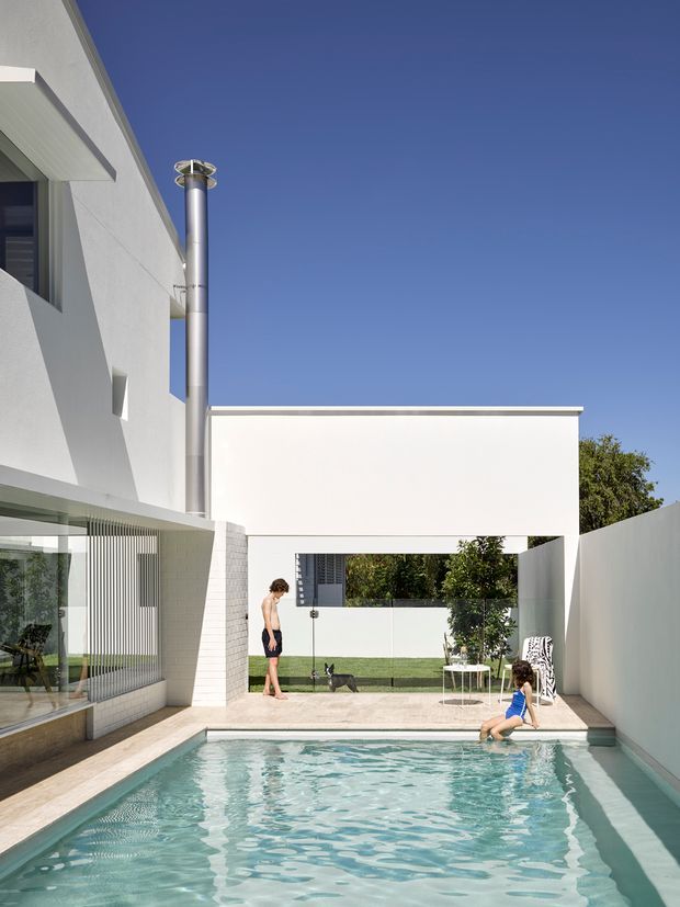 A bright contemporary residential courtyard with a rectangular swimming pool enclosed by white rendered walls. The image shows crisp geometric forms, a flat-roofed upper volume, large glazed openings with vertical screening, pale paving, and a stainless-steel chimney or flue. A small outdoor seating area and landscaped courtyard are visible beyond the pool.