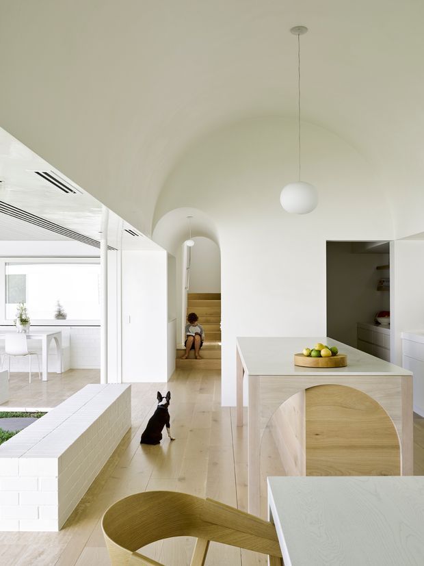 Bright, minimal open-plan residential interior with a white curved vaulted ceiling and rounded archways. The foreground is dominated by a pale timber dining table and sculptural curved-back wooden chairs, with a fruit bowl on the table. A white tiled low wall separates the dining area from a sunlit kitchen or living zone, while a staircase and built-in storage are visible in the background. The palette is predominantly white and light timber with soft natural light and clean integrated detailing.