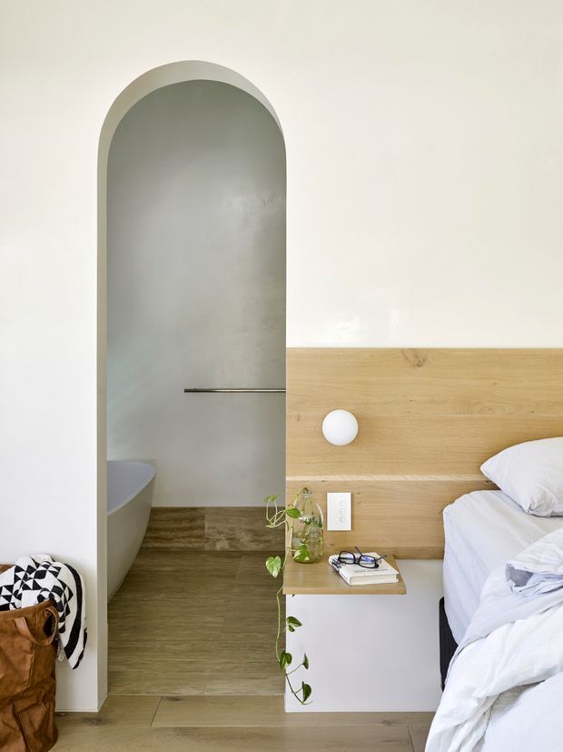 Minimalist bedroom with a white upholstered bed partially visible on the right, a full-width light timber headboard and integrated bedside shelf, and a sculptural white wall light. An arched opening leads directly to a bathroom containing a freestanding bathtub, with warm timber flooring continuing through the space. Neutral white, grey, and natural wood finishes create a calm, refined atmosphere.