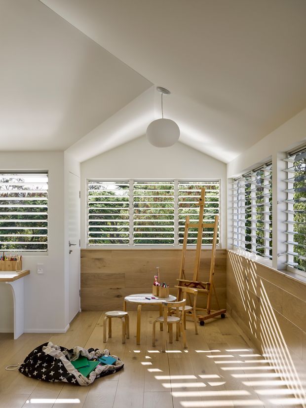 Bright, compact creative studio or children's activity room with a steep vaulted ceiling, extensive horizontal louvred windows, pale timber flooring and lower timber wall panelling. A small round table and stools, easel, art supplies and a play mat occupy the room, while strong sunlight creates striped shadows across the floor and walls.