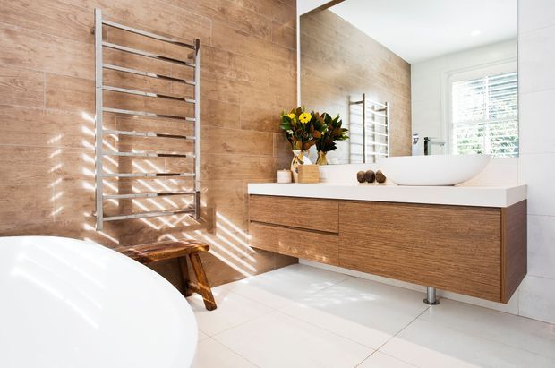 Bright contemporary bathroom featuring a freestanding white bathtub, wall-mounted timber vanity with a white benchtop and vessel basin, large mirror, white floor tiles, and a stainless-steel heated towel rail against a timber-look tiled feature wall.