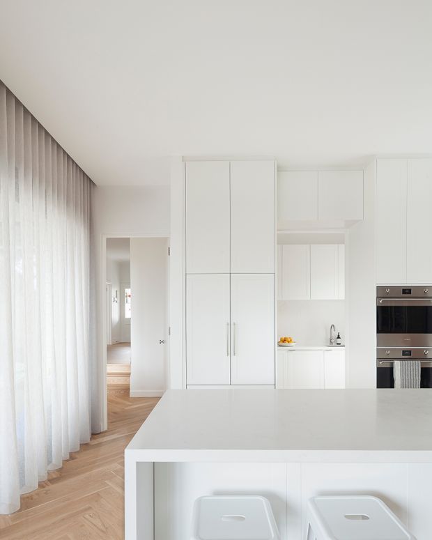 Bright, minimalist white kitchen with a large island and two white stools, full-height integrated cabinetry, built-in stainless-steel ovens, and sheer curtains along a tall window wall. Warm herringbone timber flooring extends toward an adjoining hallway.