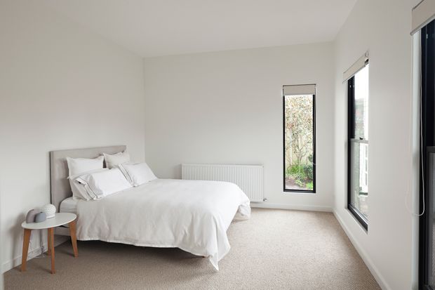Bright, minimalist bedroom with a white upholstered bed, white bedding, neutral walls, beige carpet, a small timber side table, and tall black-framed windows overlooking greenery. The room has a clean, uncluttered feel and abundant natural light.