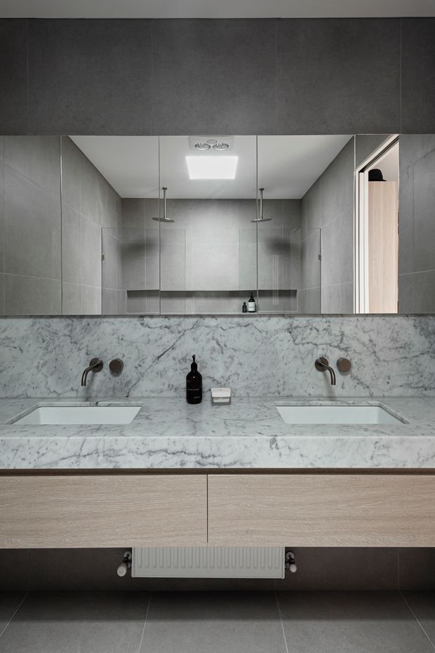 A contemporary double-vanity bathroom viewed straight on. The room features a pale veined stone vanity benchtop with two integrated rectangular basins, dark wall-mounted tapware, light timber-look drawers, large grey wall and floor tiles, a full-width mirror, and twin walk-in showers visible in the reflection. The restrained grey, white, and timber palette creates a refined, clean atmosphere.