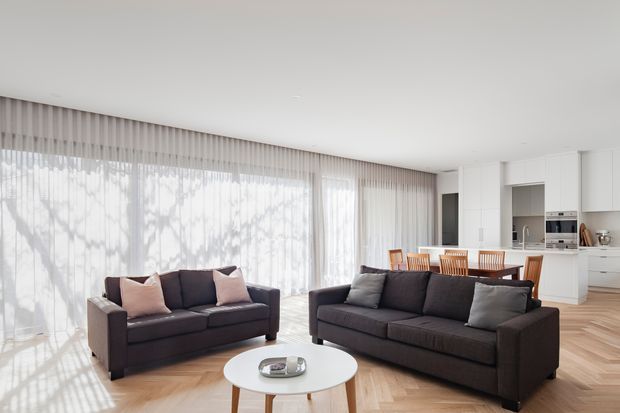Bright open-plan living room with two dark charcoal sofas, pale blush and grey cushions, a round white coffee table, full-height sheer curtains, and warm herringbone timber flooring. A white kitchen and timber dining area are visible in the background, with abundant filtered daylight creating a calm, contemporary feel.