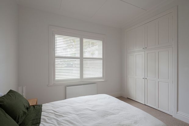 A bright, quiet bedroom with a white upholstered or covered bed in the foreground, a dark green accent cushion or throw at the left edge, white plantation-shuttered windows, a radiator, and extensive built-in white panelled wardrobes. The room has pale walls, light carpeting, simple white trim, and a clean traditional interior character.