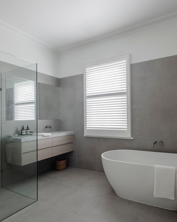 A bright, refined contemporary bathroom featuring large grey wall and floor tiles, a frameless glass shower enclosure, floating timber-look vanity with a pale stone benchtop, freestanding white bathtub, and white plantation shutters. Decorative ceiling cornices add a traditional architectural detail.