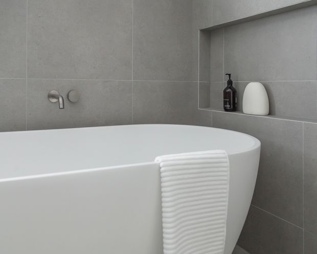 Minimal contemporary bathroom featuring a freestanding white bathtub against large-format grey tiled walls. A recessed wall niche contains dark and white bathroom accessories, while brushed metal wall-mounted tapware and a folded white ribbed towel add functional detail.