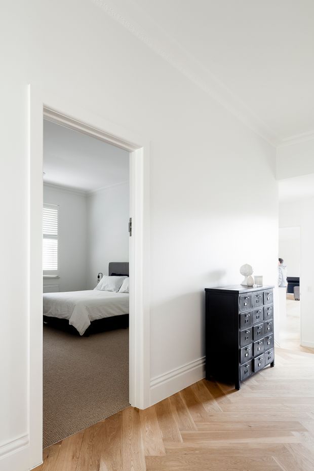 Bright, refined residential hallway with white walls, decorative trim and high ceilings, opening directly into a softly furnished bedroom. A dark chest of drawers provides contrast against the pale interior, while warm herringbone timber flooring runs through the hall and into a brighter adjoining space.