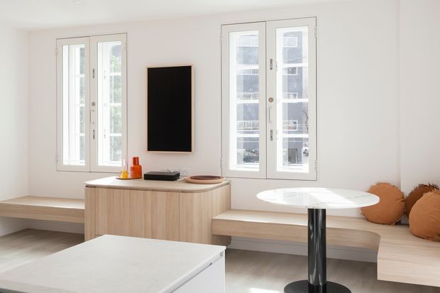 Bright minimalist dining and living nook with white walls, tall multi-pane windows, built-in light timber bench seating, a rounded light-wood sideboard, and a small round white table with a dark pedestal base. The space has clean lines, abundant natural light, and understated contemporary furnishings.