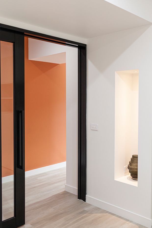 A contemporary interior hallway or entry area with pale timber-look flooring, white walls and ceiling, and a dramatic black-framed glazed sliding door opening toward a room with a vivid terracotta-orange wall. A recessed wall niche with integrated lighting displays decorative vessels. The space has clean lines, minimal detailing, and strong black, white, and orange contrast.