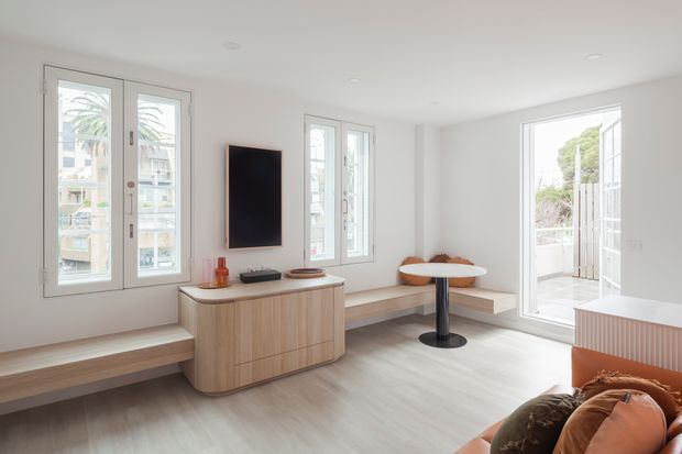 Bright, contemporary open-plan living area with white walls, pale timber flooring, large white-framed windows and glazed doors opening to a balcony or terrace. The room includes built-in timber window seating, a curved timber sideboard, a small round pedestal table, and a partial upholstered sofa in the foreground. The restrained palette and abundant daylight create an airy, calm atmosphere.
