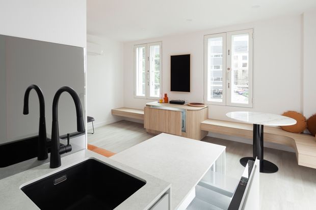 Bright open-plan contemporary apartment interior combining a compact kitchen with a living and dining area. The kitchen features a large pale stone island, matte black undermount sink and sculptural black gooseneck faucets, white cabinetry, and integrated storage. Beyond the island is a light timber built-in bench, low timber media console, white-framed windows, and a small round dining table with black pedestal base. The palette is predominantly white, light grey, pale timber, and black accents.