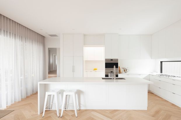 Bright contemporary open-plan kitchen with extensive white flat-panel cabinetry, a large central island with integrated sink and seating for two, integrated appliances, and light timber herringbone flooring. Sheer full-height curtains and a glazed opening create an airy, minimalist feel.
