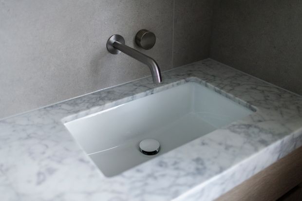 Close-up of a minimalist bathroom vanity featuring a rectangular integrated white basin set into a thick veined marble benchtop. A wall-mounted brushed metal tap and control are set against a smooth grey wall, with dark timber cabinetry visible below.