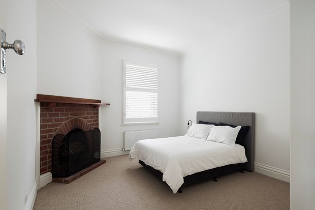 Bright, compact bedroom with a neatly made upholstered bed, white bedding, grey headboard, beige carpet, and white walls. A traditional exposed red-brick fireplace with timber mantel and black fire screen occupies the left side, while a shuttered window provides natural light.