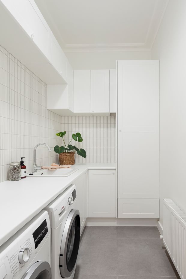Bright, compact white laundry room with a long white benchtop, integrated upper and lower cabinetry, front-loading washing machines, a deep sink with a chrome gooseneck tap, and a white vertically gridded tile backsplash. A tall built-in storage cupboard, grey tiled floor, white radiator, and a potted monstera add practical and decorative details. The room has refined moulded ceiling trim and a clean, minimalist feel.