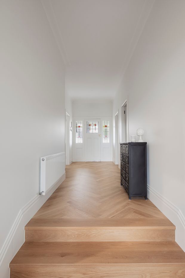 Bright, narrow residential entry hall viewed from a short flight of light timber stairs. The space has white walls and ceiling with simple decorative cornices and skirting, pale herringbone timber flooring, a white radiator, and a dark traditional chest of drawers with a spherical lamp. At the far end are white glazed entry doors with small decorative stained-glass panels.