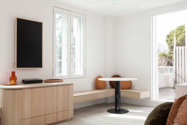 Bright, compact contemporary living and dining area with white walls, pale timber built-in bench seating, a round white tabletop on a dark pedestal base, and a light timber sideboard. Large windows and a glazed door provide abundant natural light and connect to an outdoor balcony or terrace. A wall-mounted black screen and a few warm-toned cushions and accessories add contrast.